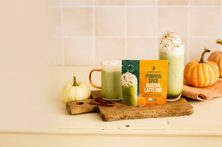 A package of Jars Leaf Pumpkin Spice Matcha Latte Mix stands on a wooden cutting board next to two prepared matcha lattes with whipped cream, with small pumpkins and cinnamon sticks in a warm kitchen setting.